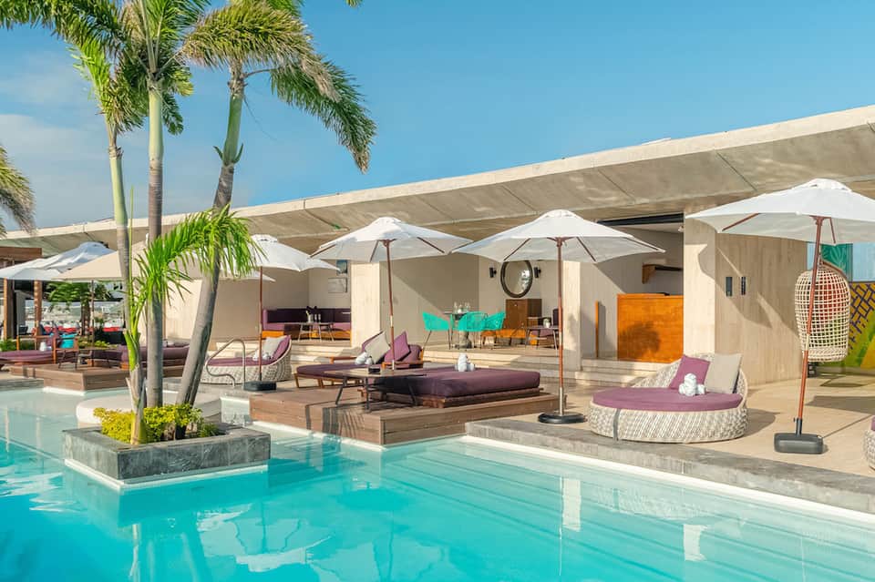 Modern pool area with white umbrellas, purple lounge chairs, palm trees, and contemporary pavilion structures