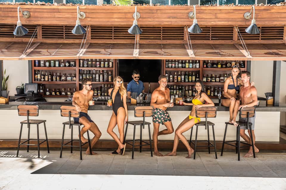 Beach bar with wooden canopy and multiple patrons in swimwear enjoying drinks