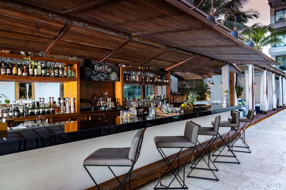 Outdoor bar with wooden pergola, black counter, bar seating, hanging plants, and tropical setting at dusk