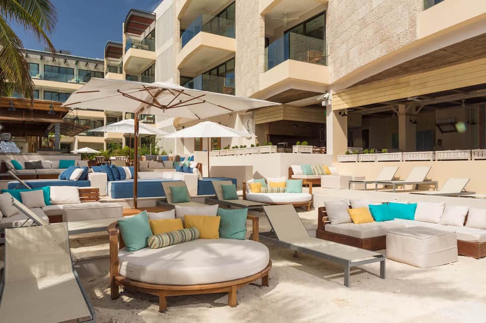 Modern beachfront resort lounge with white umbrellas, wooden loungers, and turquoise cushions