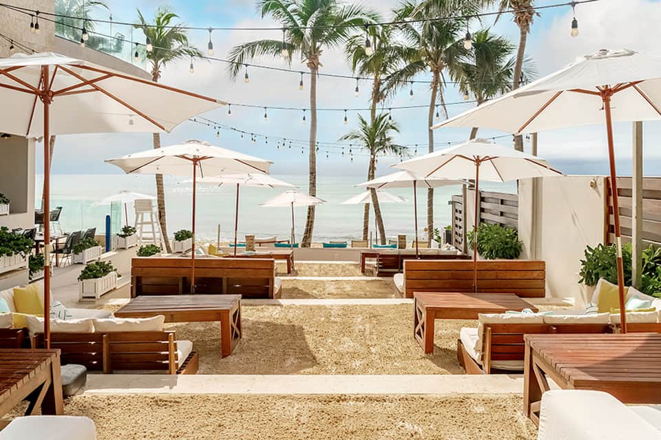 Beachfront lounge with wooden seating, cream umbrellas, palm trees, and ocean view