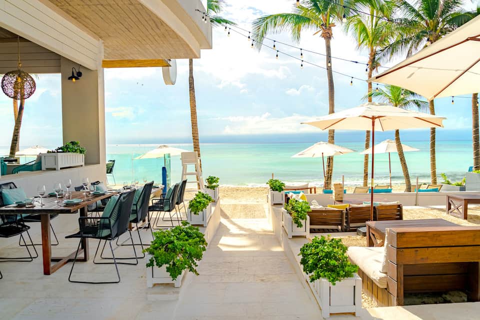 Beachfront terrace with dining tables, cream umbrellas, palm trees, and ocean views