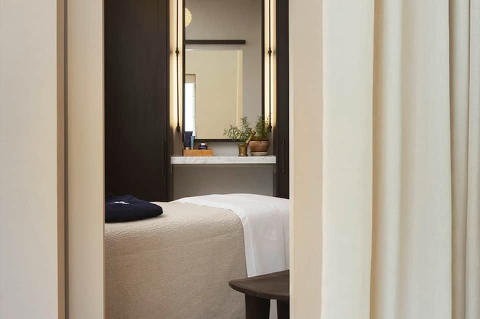 Minimalist hotel room with bed, illuminated mirror, vanity area, and neutral color palette viewed through doorway
