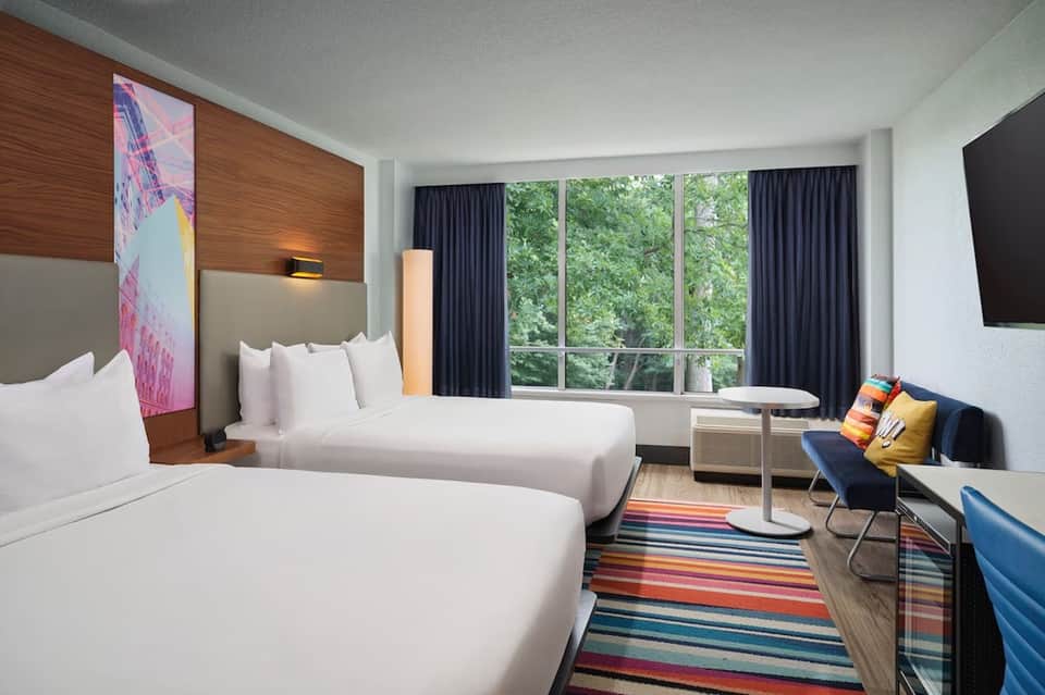 Hotel room with two queen beds, wood headboard, large windows, and colorful striped rug