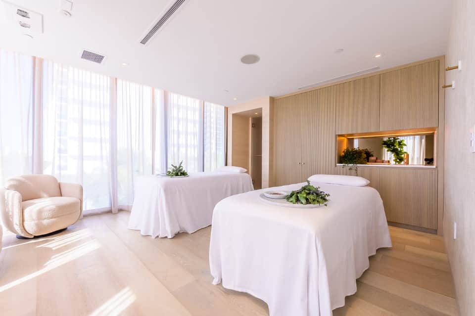 Spa treatment room with two massage tables, wooden slat accent wall, large windows, and modern lighting