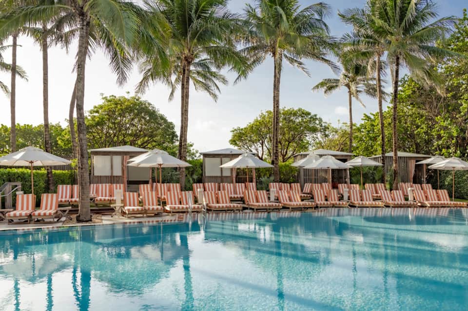 Tropical pool surrounded by palm trees with striped loungers and white umbrellas beneath clear sky
