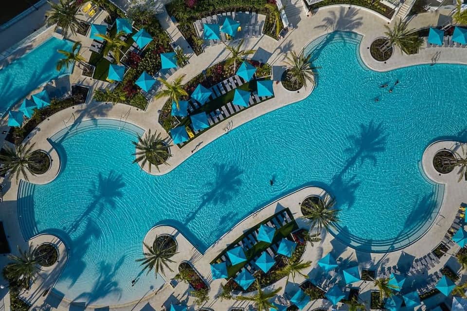 Aerial view of expansive resort pool complex with turquoise water, blue umbrellas, palm trees, and lounge chairs on surrounding deck