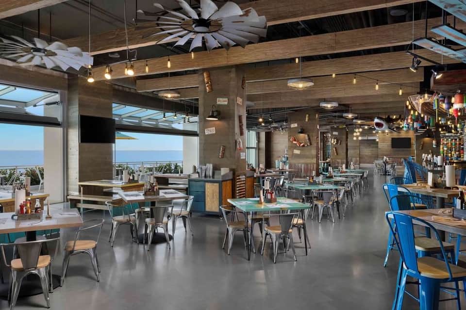 Casual beachfront restaurant with ocean views, wooden ceiling, pendant lighting, and blue accent chairs