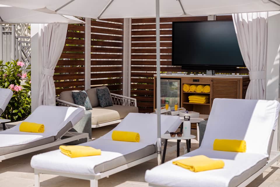 Poolside cabana with white lounge chairs, gray cushions, yellow towels, and wooden privacy screens