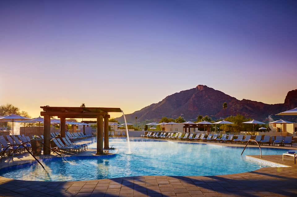 Resort pool at dusk with mountain backdrop, pergola structures, lounge chairs, and blue umbrellas