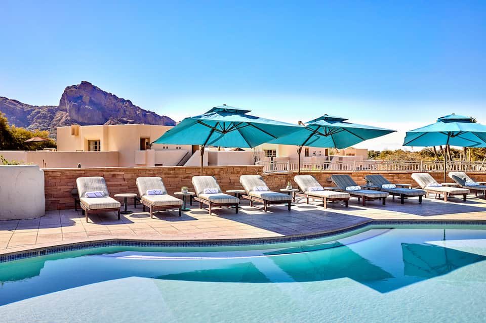 Luxury resort pool with turquoise umbrellas, white lounge chairs, mountain backdrop, and modern architecture