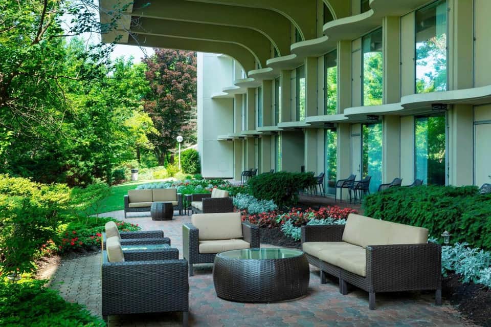 Outdoor terrace with wicker seating, round table, and lush green landscaping facing modern building