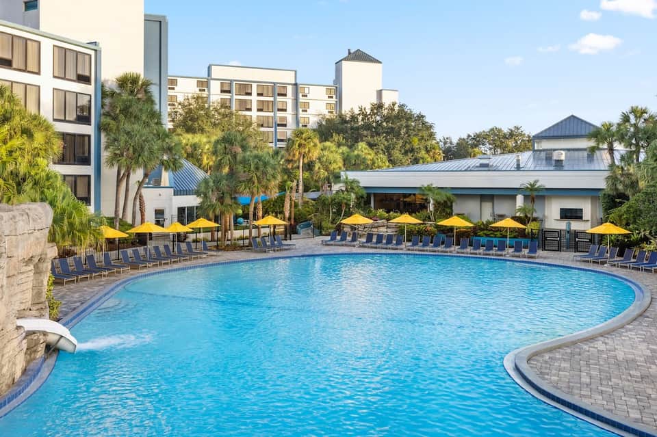 Curved resort pool with yellow umbrellas, lounge chairs, and palm trees surrounded by multi-story hotel buildings