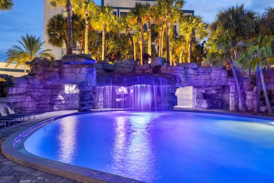 Illuminated resort pool at night with dramatic purple lighting, palm trees, and waterfall feature