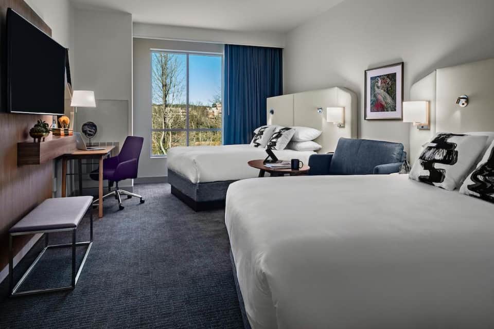 Spacious hotel room with two queen beds, desk, large windows with blue curtains, and city views