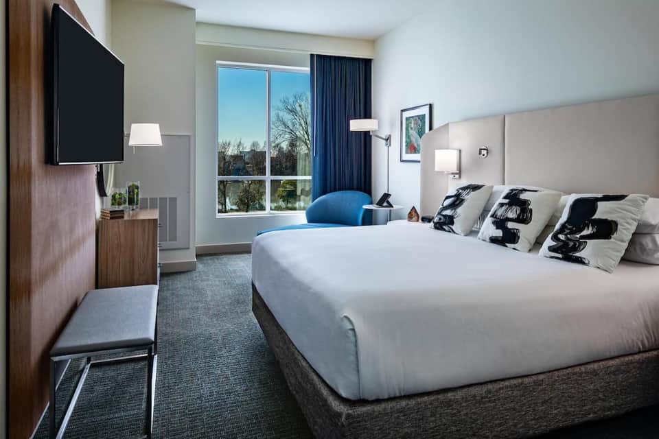 Contemporary hotel bedroom with white bedding, blue accent chair, TV, and lake view windows