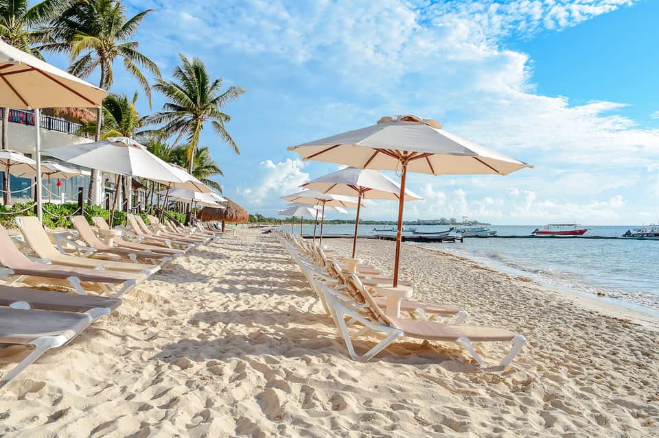 Beachfront resort with sandy shore, lounge chairs, tan umbrellas, palm trees, and moored boats in ocean