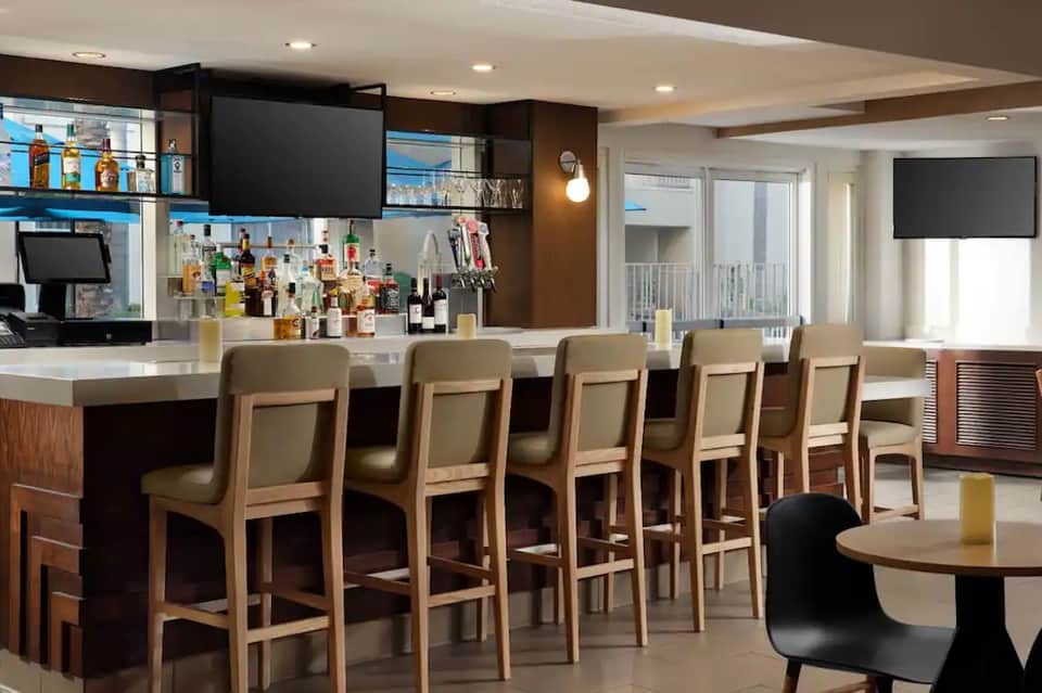 Modern hotel bar with wood accents, barstool seating, full liquor display, and flat-screen TV