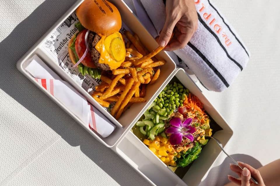 Overhead view of gourmet meal with burger, golden fries, and colorful garnished salad in takeout boxes