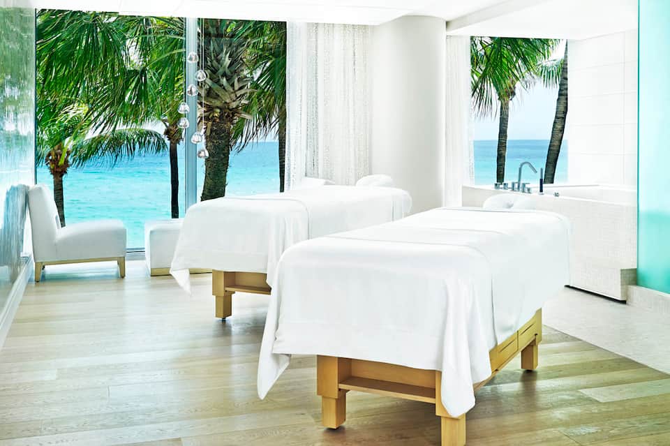 Beachfront spa treatment room with massage tables, ocean views, and palm trees through floor-to-ceiling windows