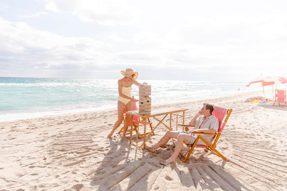Couple playing Jenga game on beach with ocean, waves, and colorful umbrellas in background