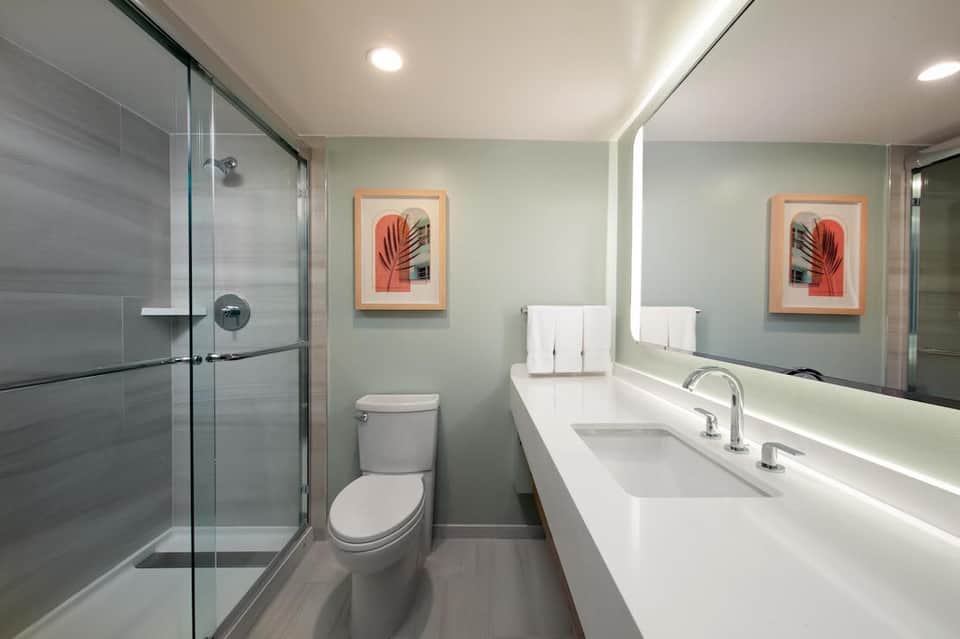 Modern hotel bathroom with mint-green walls, dual vanity sink, walk-in shower, and coral artwork