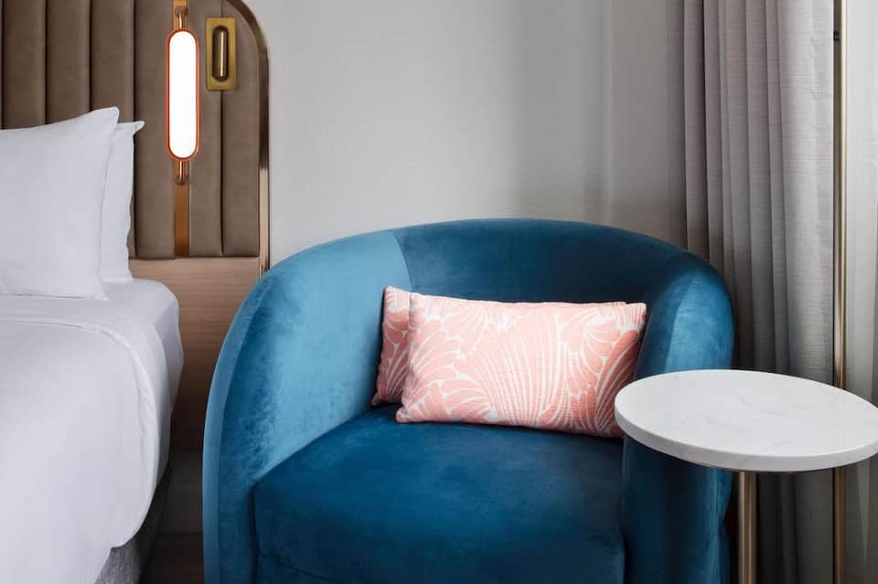 Modern hotel room with blue velvet armchair, white bed, neon wall sconce, and neutral color palette