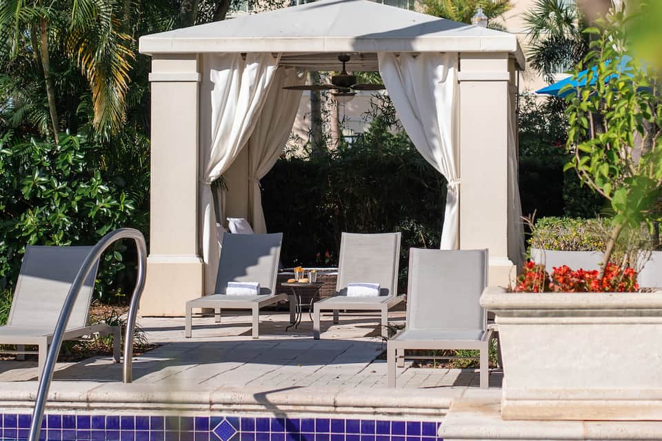 Poolside cabana with white curtains, lounge chairs, and flowering planters