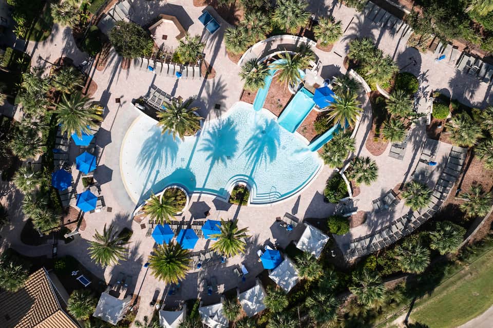 Overhead view of resort courtyard with curved pool, palm trees, blue umbrellas, and surrounding guest buildings