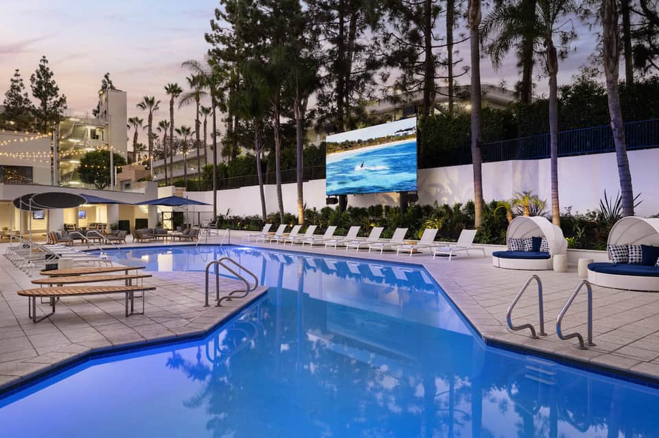 Resort pool at dusk with lounge chairs, palm trees, white cabanas, and lit buildings