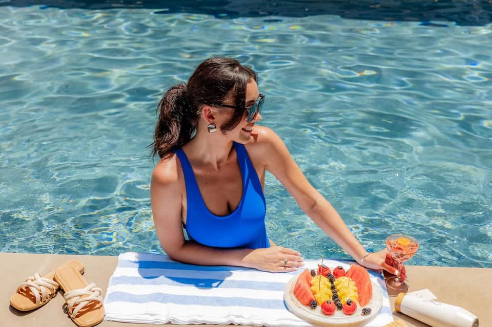 Woman in blue swimsuit relaxing poolside with fruit plate and beverage