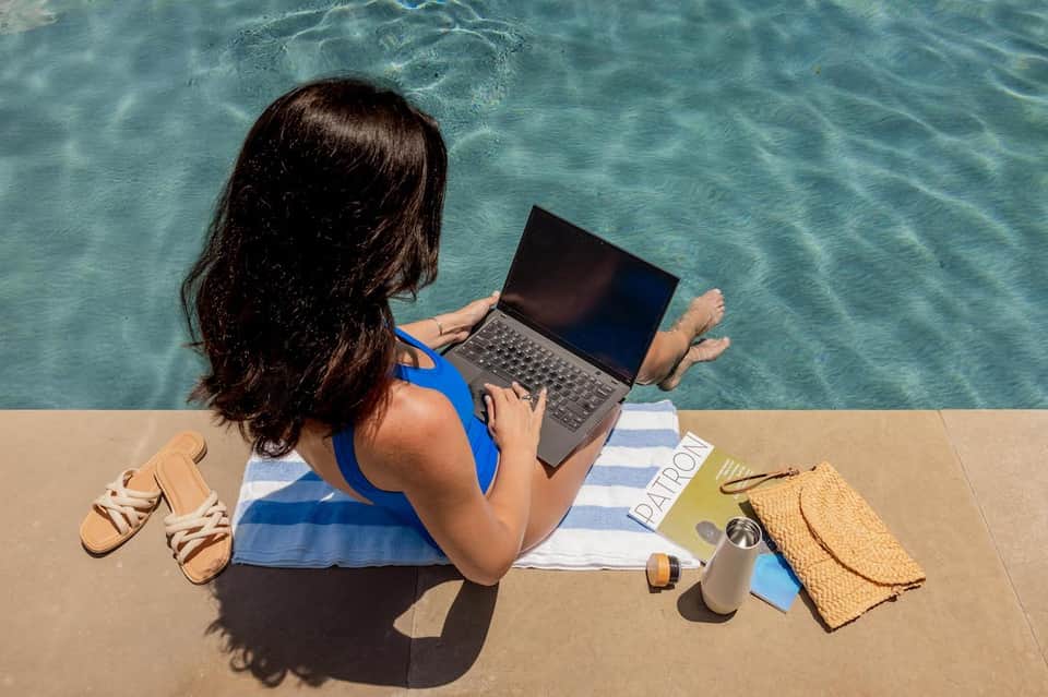Woman working on laptop poolside, sitting on striped towel in swimwear