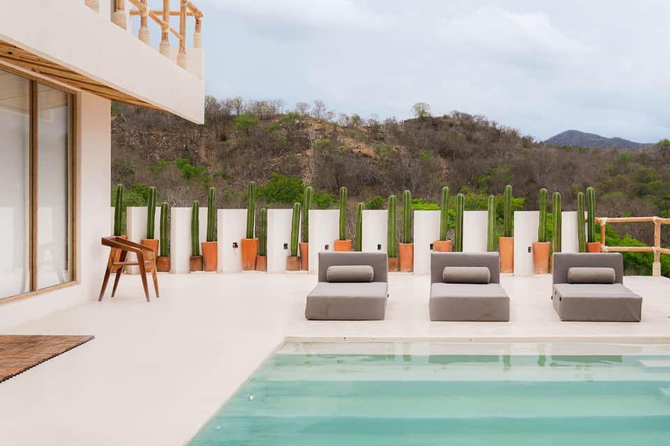 Modern patio with infinity pool, three gray lounge chairs, potted cacti, mountain views, minimalist white design