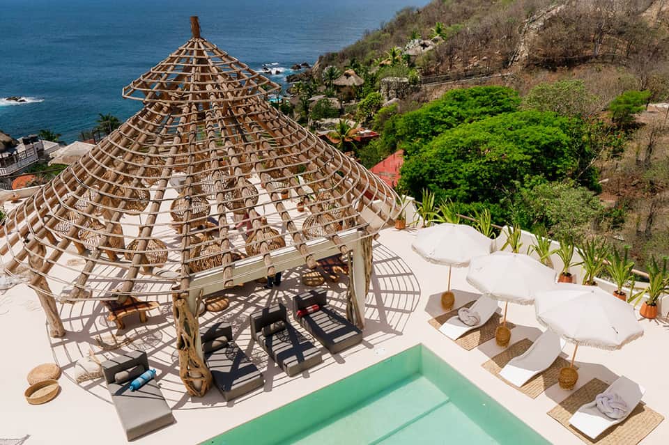 Luxury oceanside pool deck with conical woven cabana, white umbrellas, loungers, and turquoise water