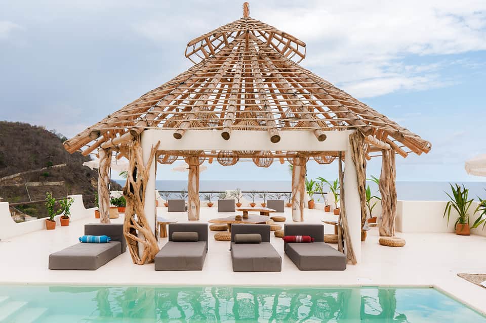 Beachfront gazebo with thatched roof, gray lounge seating, ocean view, and turquoise swimming pool
