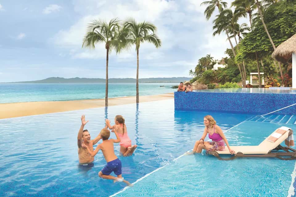 Family swimming in infinity pool with palm trees and pristine beach with ocean view