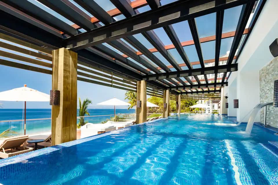 Indoor lap pool with wooden ceiling beams, ocean view, white umbrellas, and lounge chairs on the deck