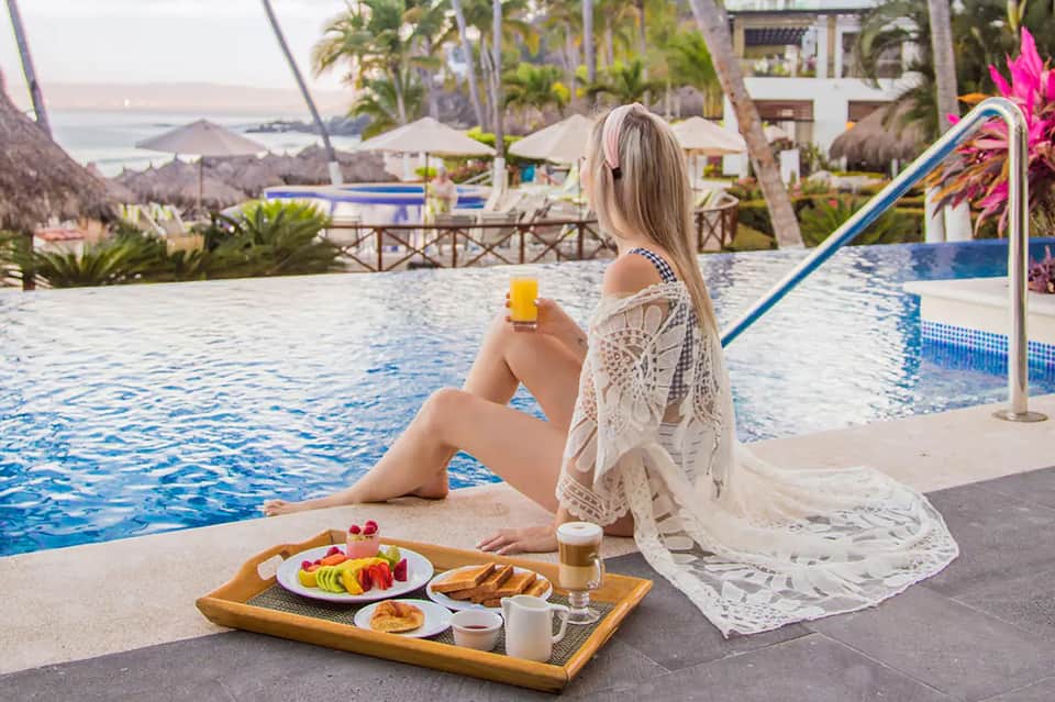 Woman relaxing poolside holding drink with breakfast tray, ocean and palm trees in background