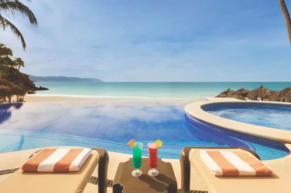 Tropical pool area with striped loungers, colorful drinks, ocean view, and palm fronds overhead