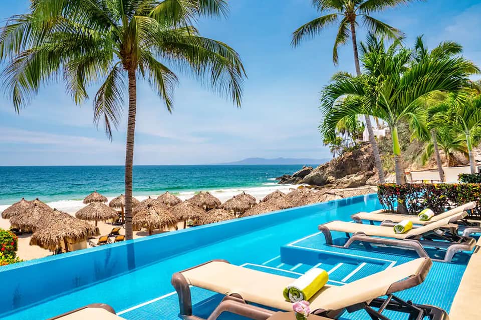 Resort pool deck overlooking tropical beach with palm trees and thatched umbrellas