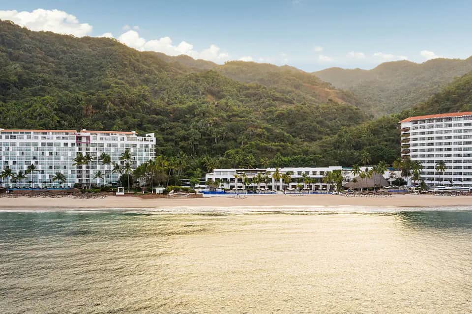 Beachfront resort with white buildings nestled against lush green mountains and sandy shoreline