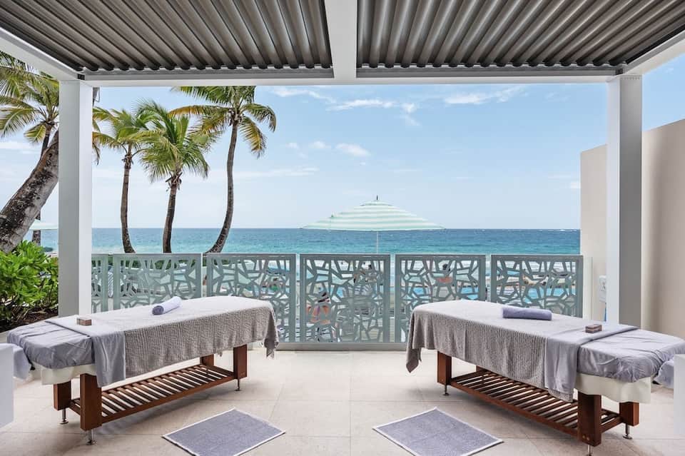 Beachfront spa treatment area with massage tables overlooking ocean, palm trees, and coastal scenery