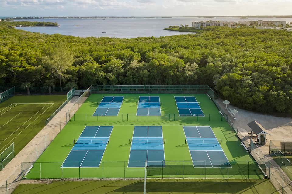 Aerial view of multiple blue tennis courts on manicured grass with waterfront and trees in background