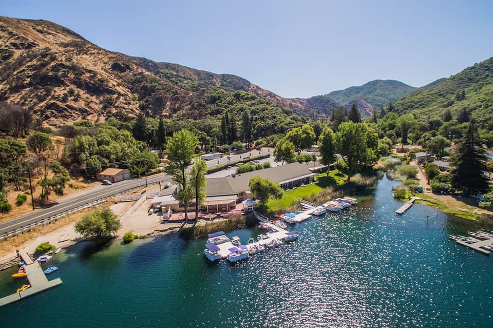 Aerial view of lakeside resort nestled in green mountains with boats, buildings, and cypress trees