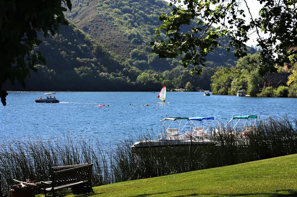 Scenic lake view with sailboat, forested mountains, and wooden bench on grassy shore