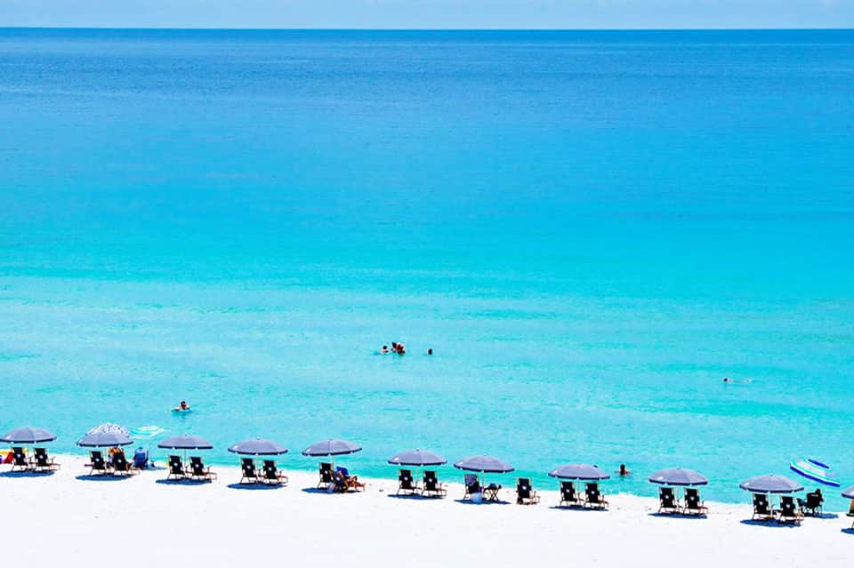 Aerial view of white sand beach lined with purple umbrellas and lounge chairs along turquoise water