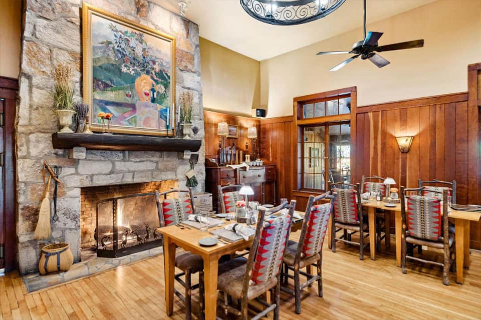 Rustic dining room with stone fireplace, wooden paneling, colorful artwork, and table set for dinner.
