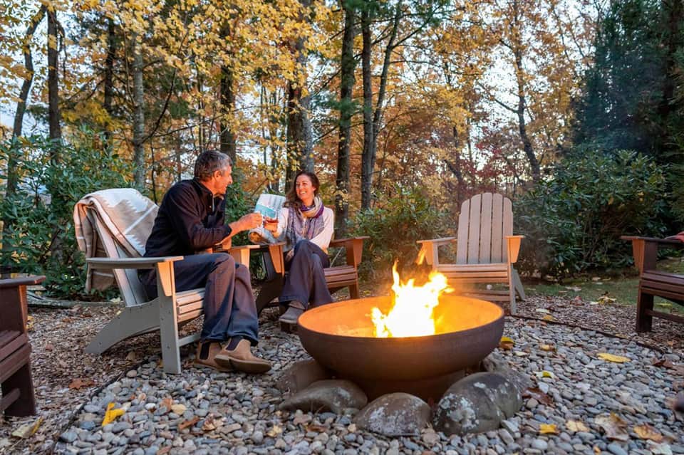 Couple sitting in Adirondack chairs around fire pit in autumn forest setting with s'mores