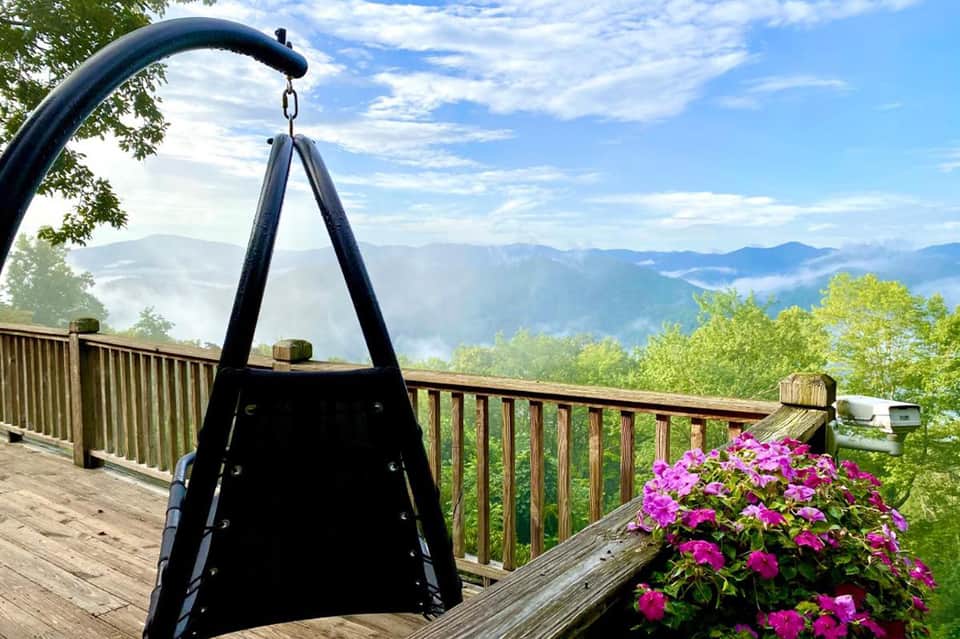 Black fire pit on wooden deck with mountain valley views, flowering magenta plants, and wooden railing overlooking misty landscape