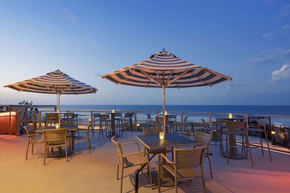 Beachfront dining area with striped umbrellas, wooden chairs, and ocean horizon at sunset